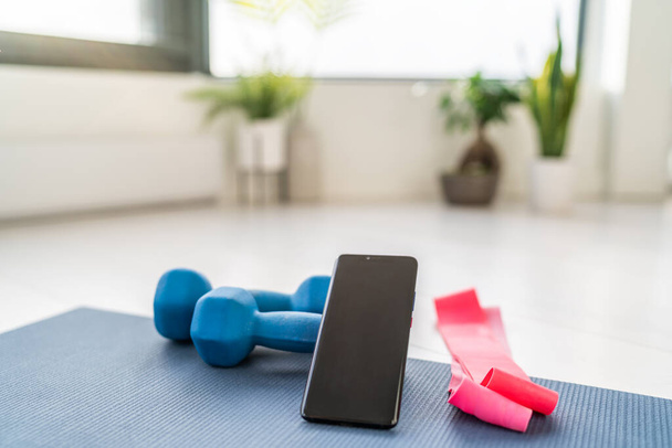 Fitness at home workout exercises online on smartphone app for training indoors at apartment with dumbbells weight and resistance bands in living room - Foto, afbeelding