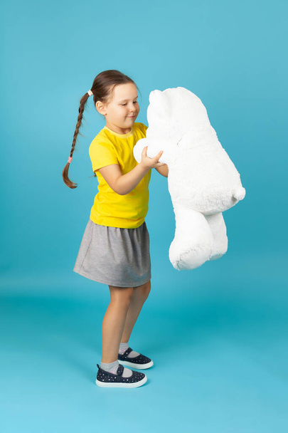 at full height the girl with pigtails in a yellow shirt and grey skirt dancing with a white Teddy bear , isolated on blue background - Φωτογραφία, εικόνα
