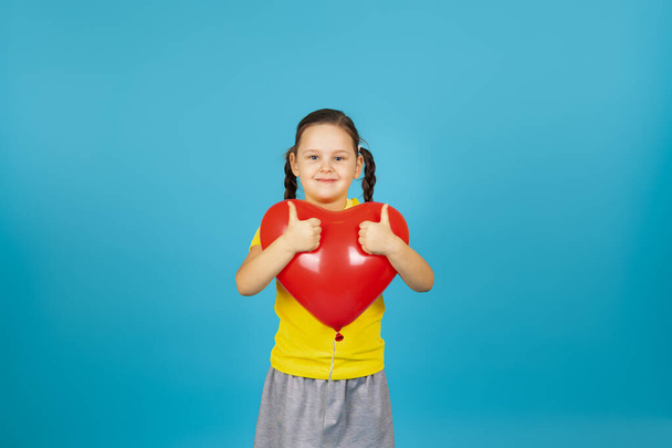 a happy child in a yellow T-shirt hugs a red heart-shaped balloon and gives a thumbs up isolated on a blue background - Foto, Imagen