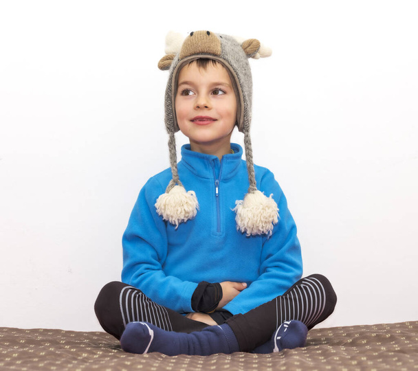 Щаслива дитина сидить схрещеними ногами на ліжку вдома, одягнена в зимовий одяг і з вовняним капелюхом у формі лося, який тримає голову теплим
. - Фото, зображення