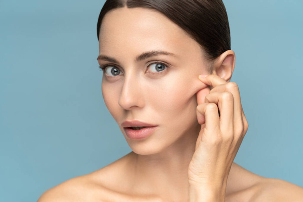 Woman without makeup touching cheeks after glycolic acid peel, has signs of aging skin on her face, looking at camera, isolated on studio blue background. Beauty skincare, cosmetology facial treatment - Photo, Image