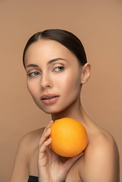 Kobieta z idealną cerą twarzy, uczesanymi włosami, trzymająca pomarańczę. Portret kobiety z naturalnym makijażem i owocami cytrusowymi na beżowym tle. Witamina C, kosmetyki kosmetyczne, koncepcja przeciwutleniająca. - Zdjęcie, obraz