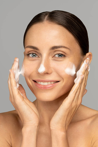 Happy young woman takes care of her skin face, applies cleansing foam after shower, smiling and looking at camera, isolated on grey background. Face wash. - Fotó, kép