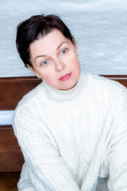 Middle Aged Woman dressed in white sweater sitting on the bedroom floor - Photo, Image