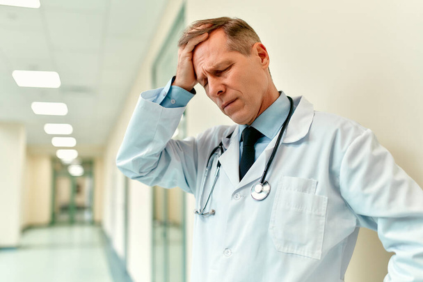 Een oudere knappe overstuur dokter in een medisch uniform met een stethoscoop staat in de gang van de kliniek en legt zijn hand op zijn hoofd als teken van wanhoop. - Foto, afbeelding