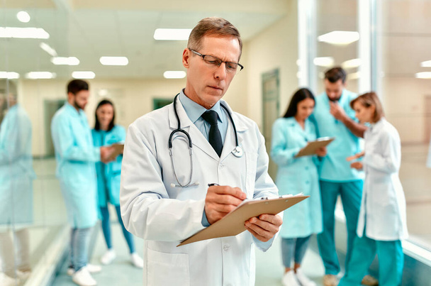 An elderly handsome serious doctor in a medical uniform with a stethoscope stands in the corridor of the clinic and makes notes against the background of the medical team. - Photo, Image