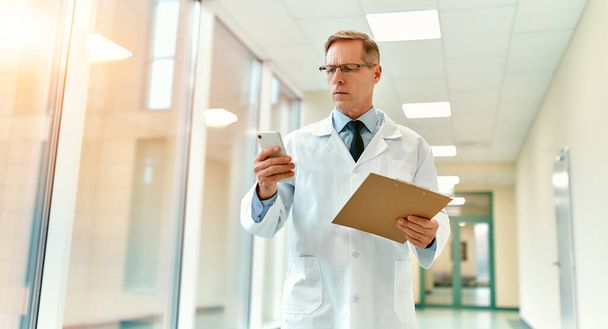 Starszy przystojny poważny lekarz w białym płaszczu i krawacie spaceruje korytarzem kliniki trzymając w ręku kartę pacjenta lub dokumenty i pisze wiadomość na smartfonie. - Zdjęcie, obraz