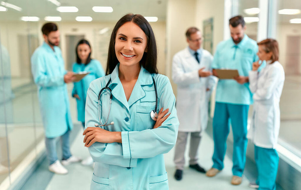 Młoda, piękna, uśmiechnięta lekarka w mundurze medycznym ze stetoskopem stoi z rękami skrzyżowanymi w korytarzu kliniki na tle zespołu medycznego. - Zdjęcie, obraz