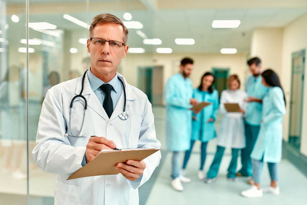 Літній красивий серйозний лікар в медичній формі зі стетоскопом стоїть в коридорі клініки і робить нотатки на тлі медичної команди
. - Фото, зображення