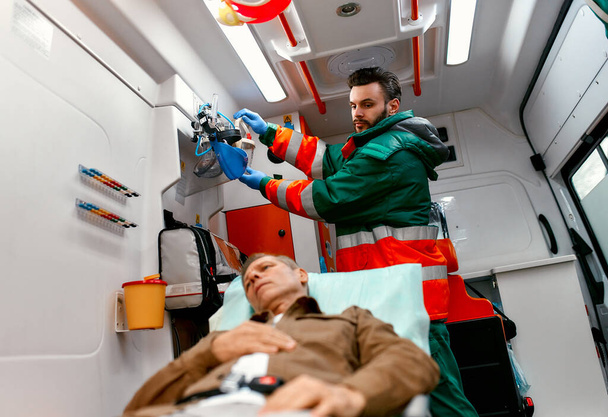 Ein männlicher Sanitäter in Uniform sucht nach einer Sauerstoffmaske, um einem älteren Patienten zu helfen, der in einem modernen Krankenwagen auf einer Liege liegt.. - Foto, Bild