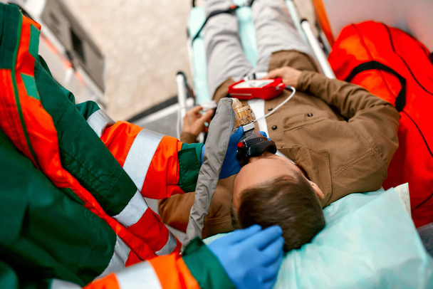 Ένας άνδρας νοσοκόμος με στολή βάζει αναπνευστήρα με οξυγόνο για να βοηθήσει έναν ηλικιωμένο ασθενή να ξαπλώσει σε ένα φορείο με ένα παλμικό οξύμετρο σε ένα σύγχρονο ασθενοφόρο.. - Φωτογραφία, εικόνα