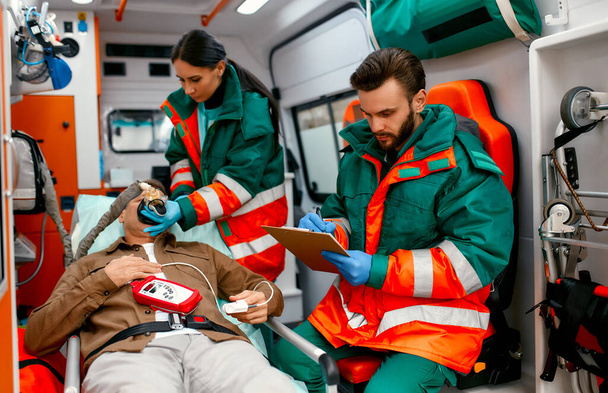 Una donna paramedica in uniforme si mette un ventilatore con ossigeno per aiutare un paziente anziano sdraiato con un pulsossimetro su una barella in un'ambulanza moderna. Un paramedico maschio guarda la cartella di un paziente. - Foto, immagini