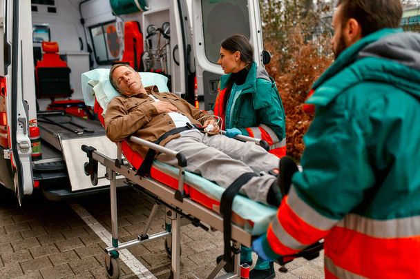 Sağlık görevlileri, klinikte tıbbi bakım için modern bir ambulanstan yaşlı bir hastayla birlikte sedye çıkarıyorlar.. - Fotoğraf, Görsel