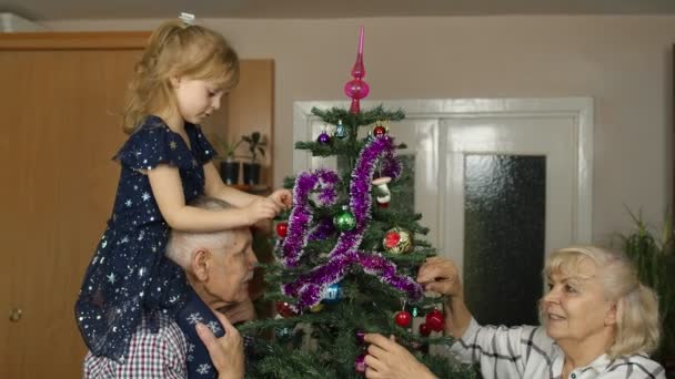 Маленькая милая девочка со старшими бабушкой и дедушкой украшают искусственную елку дома - Кадры, видео