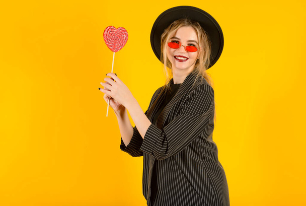 1 young blonde woman in black suit, hat with red heart on yellow background - Фото, зображення