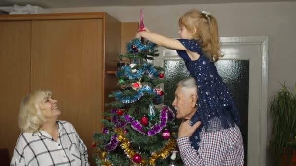 Маленькая милая девочка со старшими бабушкой и дедушкой украшают искусственную елку дома - Кадры, видео