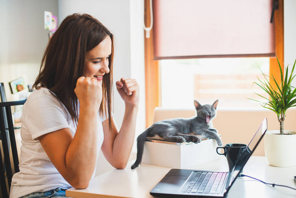 Jonge vrouw in wit t-shirt zittend met een kat op schoot aan de houten tafel thuis met laptop en notebook, aan het werk - Foto, afbeelding