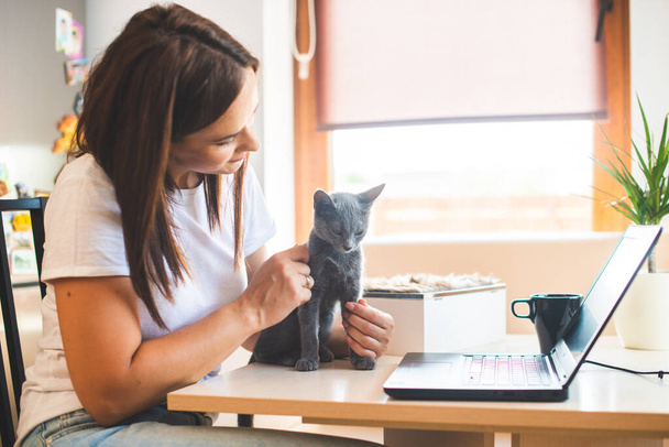 Junge Frau im weißen T-Shirt sitzt mit Katze auf dem Schoß am Holztisch zu Hause mit Laptop und Notizbuch und arbeitet - Foto, Bild