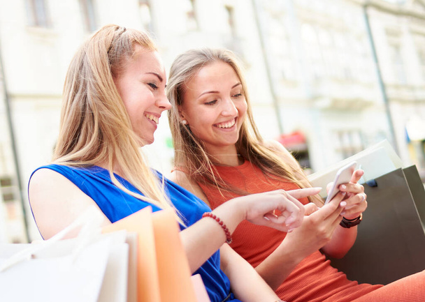 Молоді тисячолітні жінки використовують смартфон для онлайн-покупки - Модні дівчата сидять на лавці, серфінг в Інтернеті з мобільними телефонами - концепція продажів та веб-покупців
 - Фото, зображення