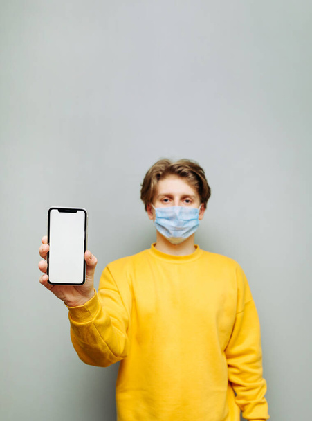 The guy in casual clothes stands on a background of a gray wall with a smartphone in his hands and shows a copy space screen with a serious face. - Photo, Image
