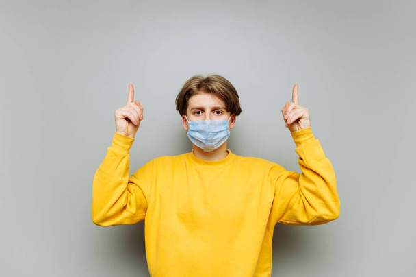 Handsome young man in a protective mask shows thumbs up on a blank space and looks at the camera on a background of gray wall. Coronavirus concept. - Photo, Image