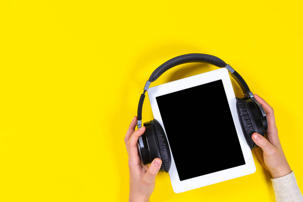 Online learning, remote education, e-learning concept. Kid hands holding digital tablet with wireless headphones over yellow background. Top view - Photo, Image