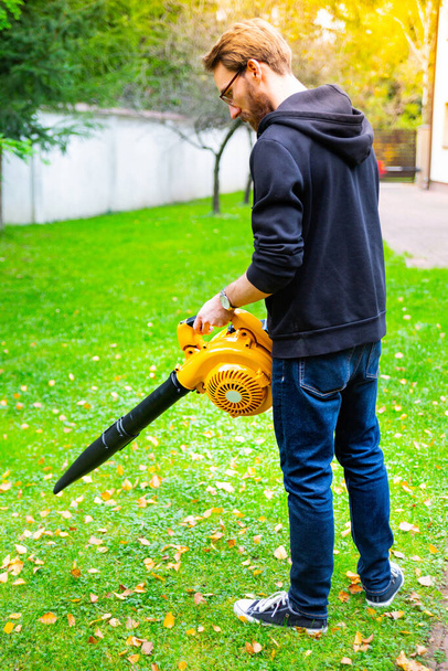 A young bearded man using a cordless, electric leaf blower in a garden. Garden works. Autumn, fall gardening works in a backyard, on a lawn, grass. - Фото, изображение