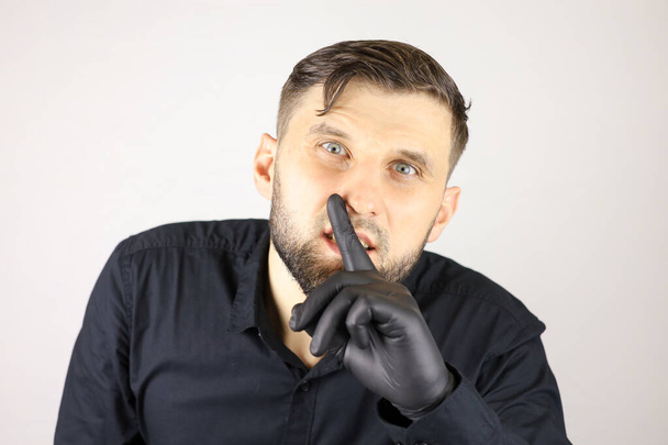 Ένας άντρας που φοράει ιατρικά γάντια βάζει το δείκτη του στα χείλη του και κάνει μια ήσυχη χειρονομία - Φωτογραφία, εικόνα
