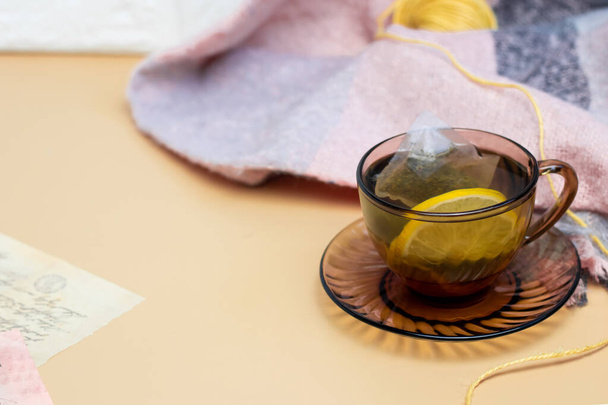 Green tea with lemon in a transparent dark mug on a yellow background and a knitted scarf. Cozy wallpapers, Home atmosphere, Copy Space - Photo, Image