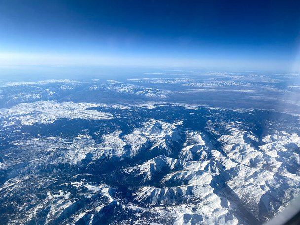 Una vista aérea desde una ventana de avión de montañas, nieve, nubes y cielos azules brillantes. - Foto, Imagen