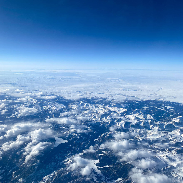 Vue aérienne depuis une fenêtre d'avion de montagnes, neige, nuages et ciel bleu vif. - Photo, image