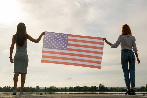 Silueta de dos jóvenes amigas que sostienen la bandera nacional de Estados Unidos en sus manos paradas juntas al aire libre. Chicas patrióticas celebrando el Día de la Independencia de los Estados Unidos. Día Internacional de la Democracia. - Foto, imagen