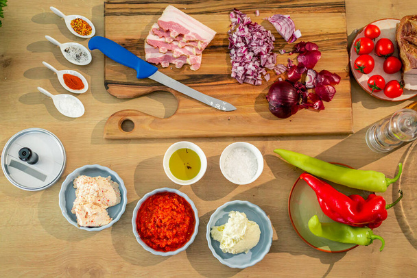 Vista superior de la superficie de trabajo de la cocina con tocino, cuchillo y comestibles, como especias y verduras en una tabla de madera. - Foto, Imagen