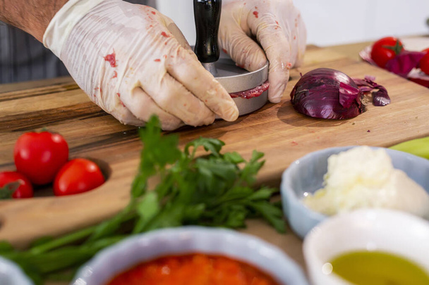 Κοντινό πλάνο ενός σεφ που φτιάχνει κρέας για μπιφτέκια σε πιεστήριο σε ξύλινη σανίδα στην κουζίνα. Υπάρχουν διάφορα μπαχαρικά, λαχανικά, καθώς και μικρά μπολ κουζίνας γύρω από την επιφάνεια εργασίας. - Φωτογραφία, εικόνα