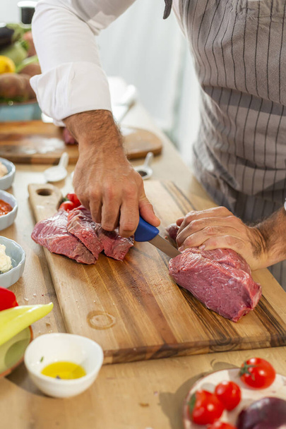 Un primo piano delle mani dello chef che taglia la carne a pezzi sottili con un coltello. Diverse spezie e verdure possono essere visti intorno. - Foto, immagini