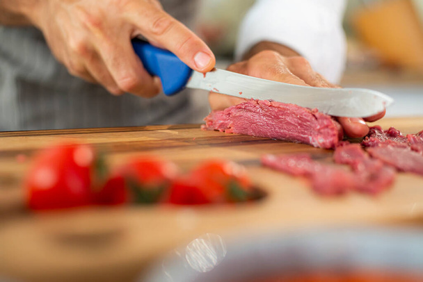 Um close-up das mãos do chef cortando carne de carpaccio em pedaços finos durante a fase de preparação em uma placa de cozinha de madeira. Várias pequenas tigelas de cozinha e legumes podem ser vistos ao redor. - Foto, Imagem