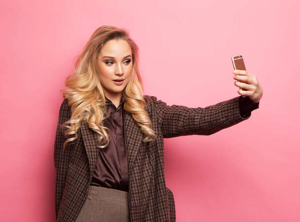 Portrait of smiling blond woman in casual clothes with perfect makeup and hair style with smartphone, makes selfie over pink background. Fashion and people concept. - Φωτογραφία, εικόνα