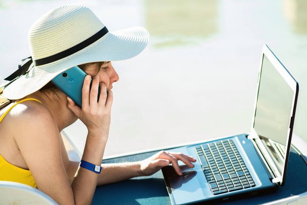 Nuori nainen rannalla tuoli uima-altaalla työskentelee tietokoneen kannettava ja puhuu myydä puhelimeen kesällä keinona. Etätyö ja freelance työtä matkoilla käsite. - Valokuva, kuva