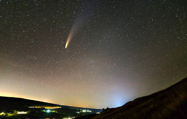 Noche de verano en montañas con cielo nublado azul oscuro estrellado y cometa C / 2020 F3 (NEOWISE) con cola clara y luces brillantes del pueblo. - Foto, imagen