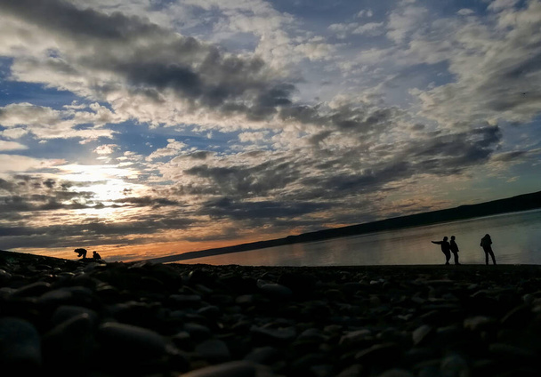 prachtig landschap zonsondergang met silhouetten onherkenbare mensen, van vijf personen in totaal, drie bij het water of de kust en een wijzen, met bergen op de achtergrond en rondom ondoorzichtige Toon - Foto, afbeelding
