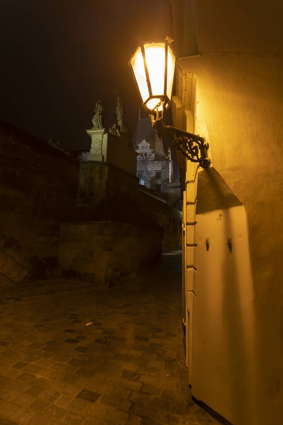 Prag 'ın merkezinde kaldırım taşları olan karanlık bir caddedeki sokak lambasının ışığı ve Çek Cumhuriyeti' nin Prag 'ın merkezinde Charles Köprüsü' ndeki taş köprü kulesi. - Fotoğraf, Görsel