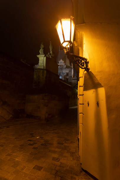lumière d'un lampadaire dans une rue sombre avec pavés dans le centre de la prague et une tour de pont en pierre sur le pont Charles dans le centre de la prague dans la république tchèque - Photo, image