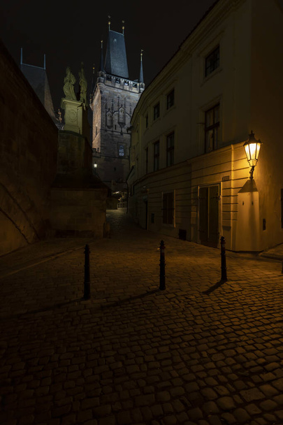Prag 'ın merkezinde kaldırım taşları olan karanlık bir caddedeki sokak lambasının ışığı ve Çek Cumhuriyeti' nin Prag 'ın merkezinde Charles Köprüsü' ndeki taş köprü kulesi. - Fotoğraf, Görsel