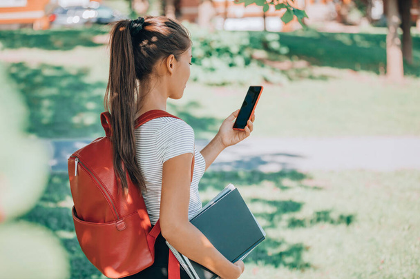 Back to school semester online on phone app. University student using mobile phone for watching her classes while walking on campus with backpack, laptop, book. Woman holding cellphone - Zdjęcie, obraz