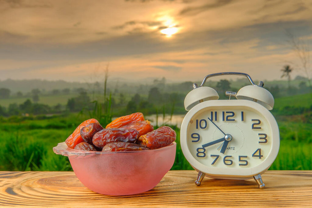  Fechas secas fruta en un tazón y despertador en madera vieja con un hermoso amanecer como fondo. El mes santo del concepto de Ramadán. - Foto, imagen