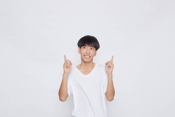 Muotokuva iloinen hymyilevä aasialainen mies yllään rento valkoinen t-paita osoittaa käsi tyhjään tilaan vieressä valkoisella pohjalla - Valokuva, kuva