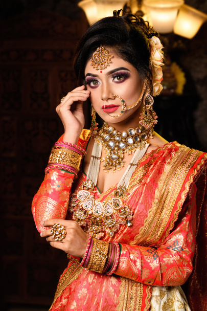 Portret van een zeer mooie jonge Indiase bruid in luxe bruidskostuum met make-up en zware sieraden in studio verlichting indoor - Foto, afbeelding