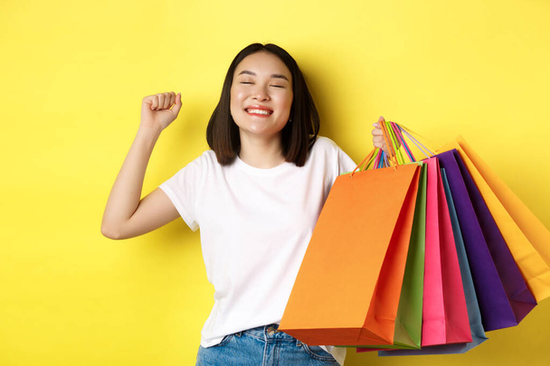 Szczęśliwa azjatycka kobieta czuje się zadowolona po zakupach na sprzedaż, trzymając papierowe torby i rozciągając się z uśmiechem, stojąc na żółtym tle - Zdjęcie, obraz