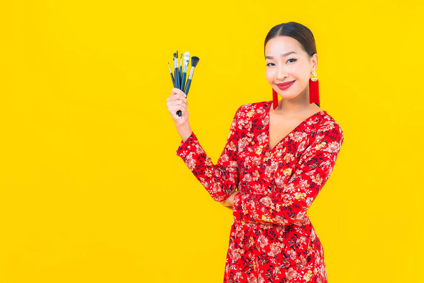 Ritratto bella giovane donna asiatica con make up pennello e cosmetici su sfondo giallo - Foto, immagini