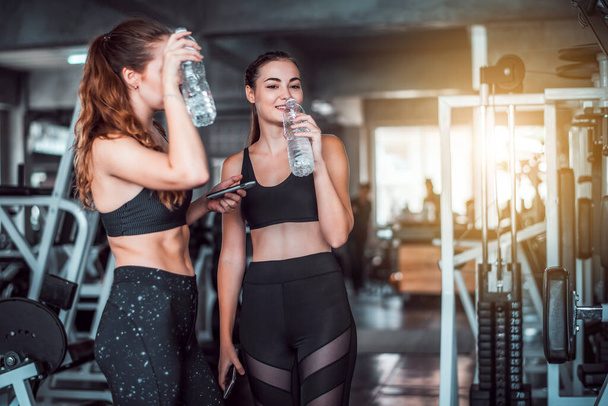Dwie młode, atrakcyjne, kaukaskie kobiety rozmawiające ze sobą i korzystające ze smartfona lub telefonu komórkowego podczas picia wody i relaksu po ciężkim treningu na siłowni  - Zdjęcie, obraz